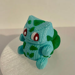 Handmade knitted-style 3D printed Pokémon-inspired plant creature with textured filament, large eyes, teal body and green accents, photographed from the side on a white background.