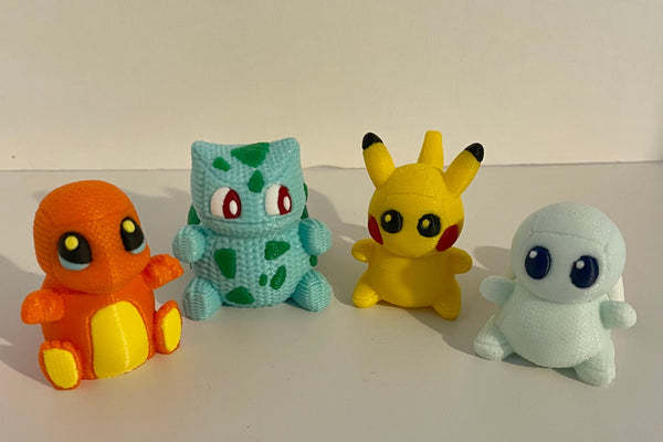 Four handmade knitted-style 3D printed Pokémon-inspired collectibles featuring textured filament, bright colors, and cute chibi designs, including an orange fire character, green plant dinosaur, yellow electric mouse, and light blue turtle displayed in a row on a white background.